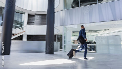 Businessman rushing, pulling suitcase in modern office lobby