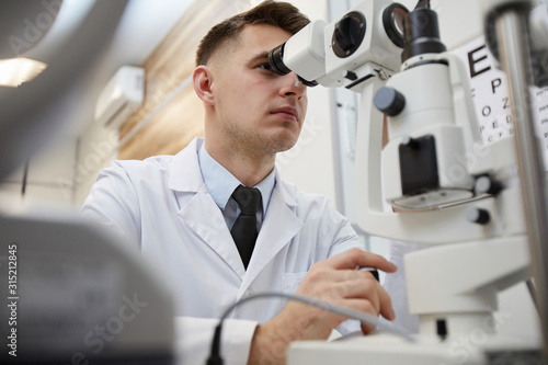 Low angle portrait of male optometrist using refractometer machine while testing vision of unrecognizable patient
