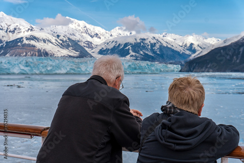 Old couple on a cruise ship in Alaska. Husband and wife are enjoying an amazing view of Hubbard Glacier and iceberg. Snow peaks and ice all around in the sea.