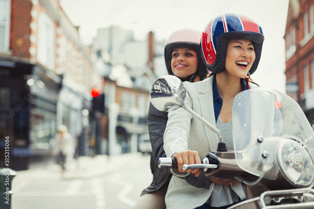 Smiling young women friends wearing helmets riding motor scooter on urban street Smiling young women friends wearing helmets riding motor scooter on urban street