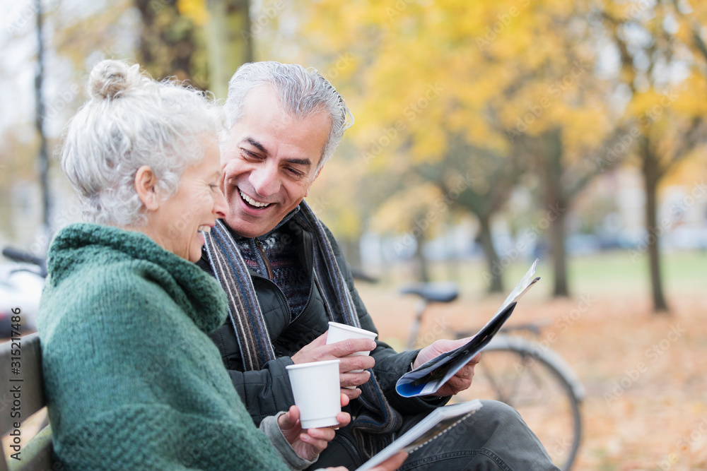Smiling senior couple reading newspaper and drinking coffee on bench in autumn park Smiling senior couple reading newspaper and drinking coffee on bench in autumn park