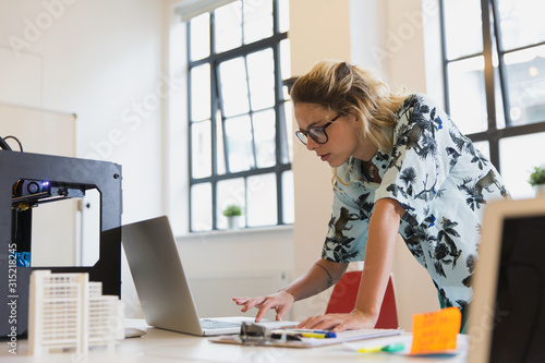 Female designer working at laptop next to 3D printer in office photo