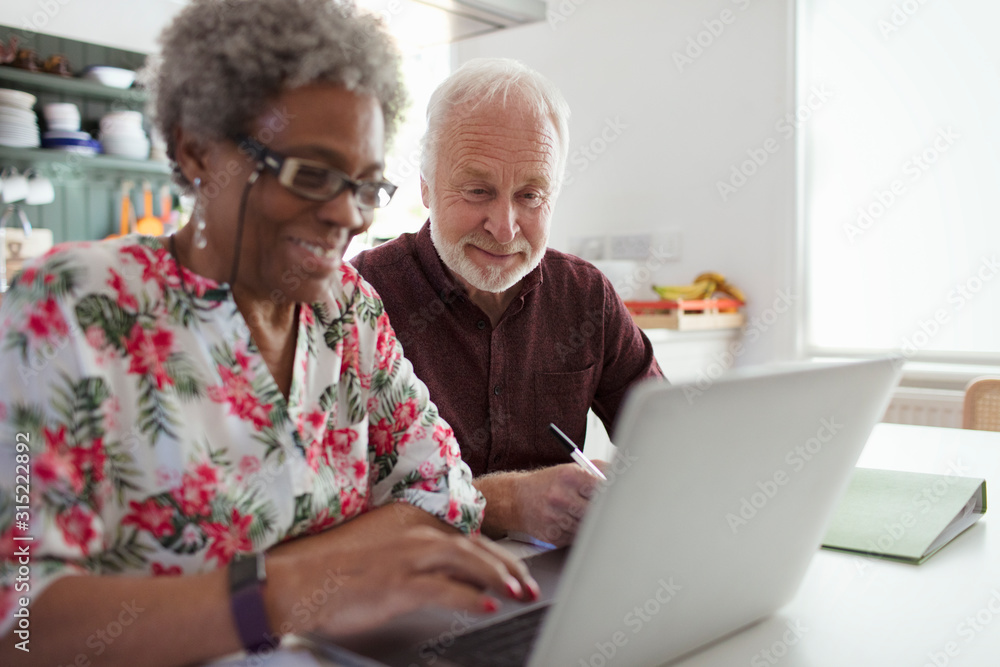 Senior couple paying bills at laptop in kitchen Senior couple paying bills at laptop in kitchen