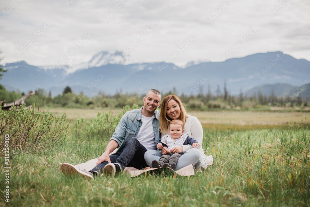 Portrait smiling parents baby son sitting in rural field mountains Portrait smiling parents baby son sitting in rural field mountains