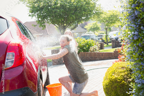 Wallpaper Mural Playful daughter spraying mother hose, washing car in sunny driveway Torontodigital.ca