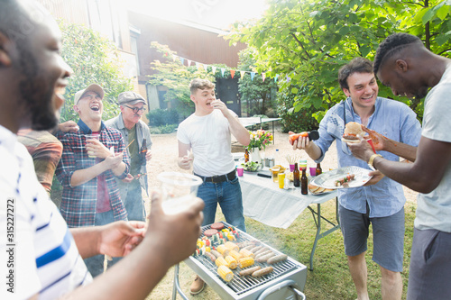 Male friends laughing and eating around barbecue grill in backyard photo
