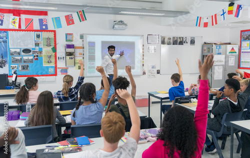 Male teacher leading lesson at projection screen in classroom students raising hands