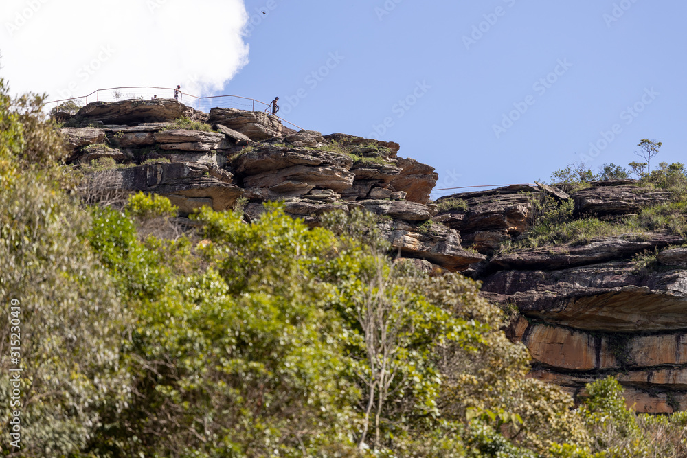 Naklejka premium People as small silhoutted specks on the huge plateau with sticking out rock formations in the Andorinhas [swallows] park in Ouro Preto seen from a lower viewpoint against a blue sky with cloud