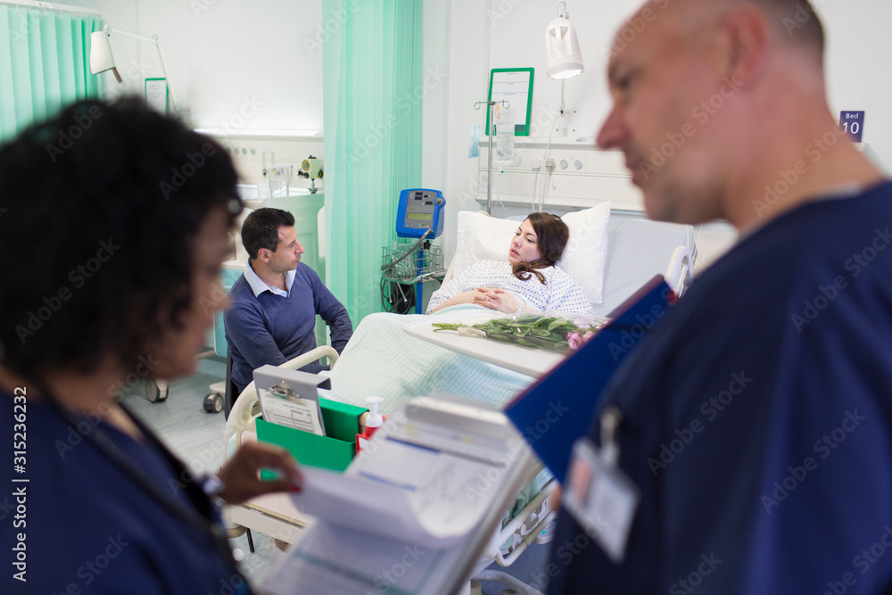 © Tom Merton/Caia Image - Doctors with medical chart making rounds with patient resting in hospital bed in background © Tom Merton/Caia Image - Doctors with medical chart making rounds with patient resting in hospital bed in background