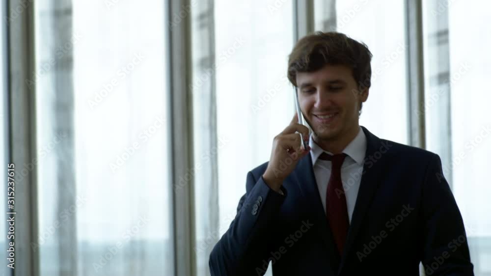 Medium shot with slowmo of successful businessman in suit talking to business partner on phone and walking along hall of office building with panoramic windows Medium shot with slowmo of successful businessman in suit talking to business partner on phone and walking along hall of office building with panoramic windows