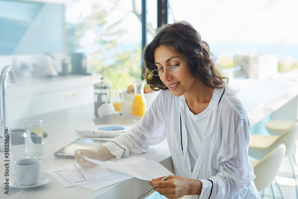 Businesswoman in bathrobe working reviewing paperwork at kitchen counter Businesswoman in bathrobe working reviewing paperwork at kitchen counter