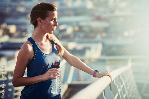 Female runner with water bottle resting on sunny urban footbridge