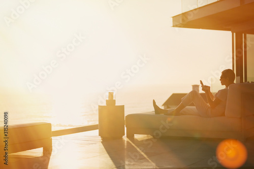 Silhouette woman using cell phone on chaise lounge on luxury balcony with sunset ocean view