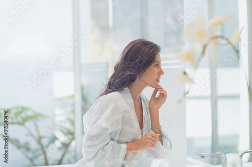 Brunette woman in bathrobe touching chin at bathroom mirror photo