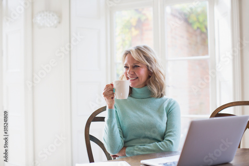 Smiling mature female freelancer drinking tea and working at laptop at home