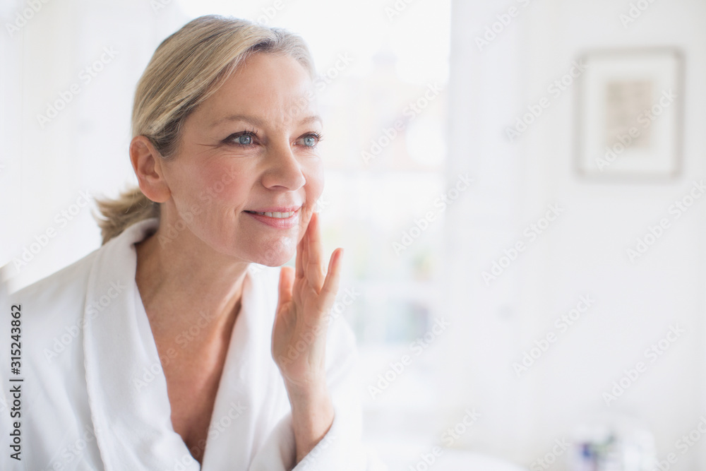 Smiling mature woman applying moisturizer to face at bathroom mirror Smiling mature woman applying moisturizer to face at bathroom mirror