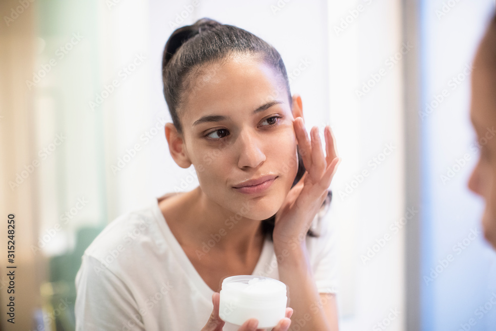 Woman applying moisturizer to face in bathroom mirror Woman applying moisturizer to face in bathroom mirror