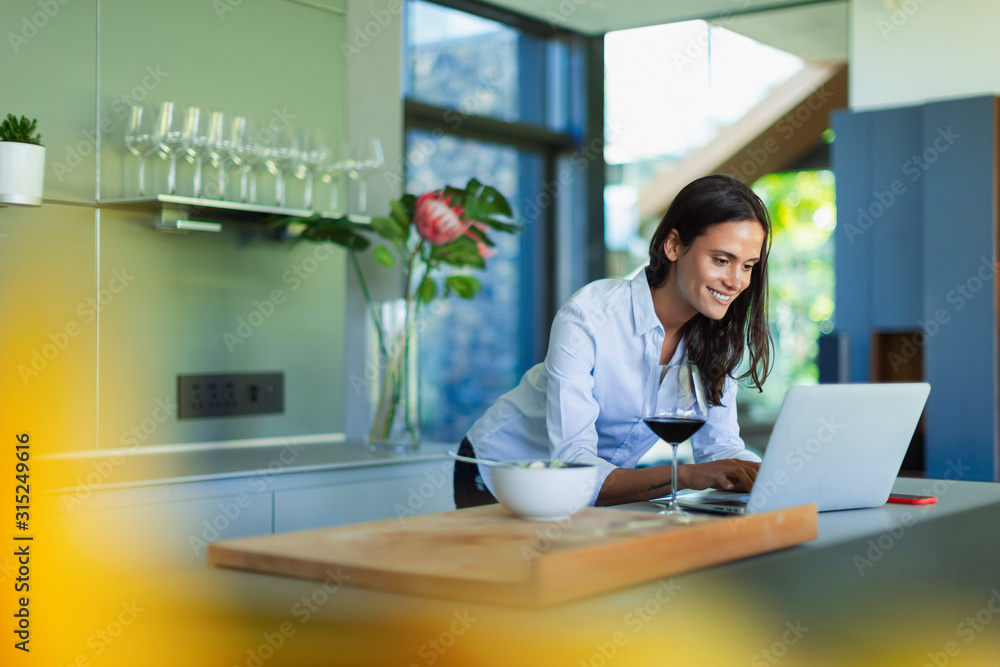 Smiling woman using laptop and drinking red wine in kitchen Smiling woman using laptop and drinking red wine in kitchen