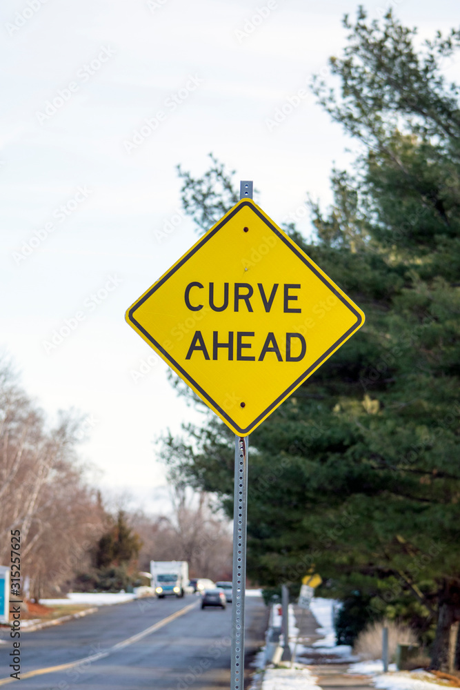 A road sign CURVE AHEAD made in a diamond shape painted in yellow and black colors attached to a signpost.