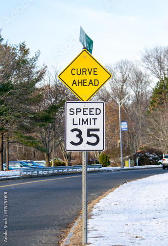 A road sign CURVE AHEAD made in a diamond shape combined with SPEED LIMIT sign, painted in yellow and black colors attached to a signpost.