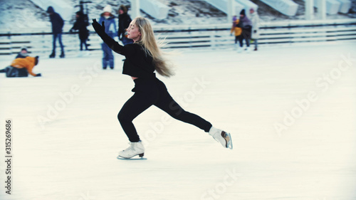 A young blonde attractive woman figure skater skating on the outdoors ice rink around people