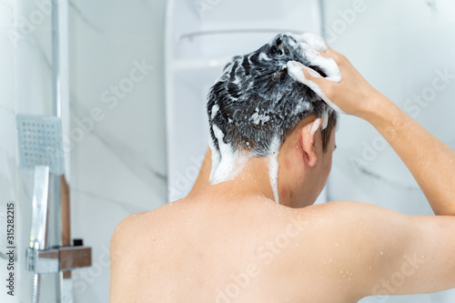 Back view from below of black haired naked man washing hair with shampoo while talking shower in bathroom