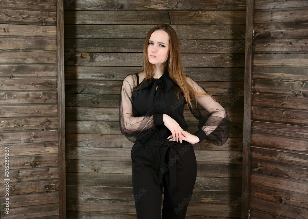 Fototapeta premium The concept of home comfort. Photo of a pretty brunette girl with excellent make-up in dark clothes standing on a wooden background in the home interior, smiling.