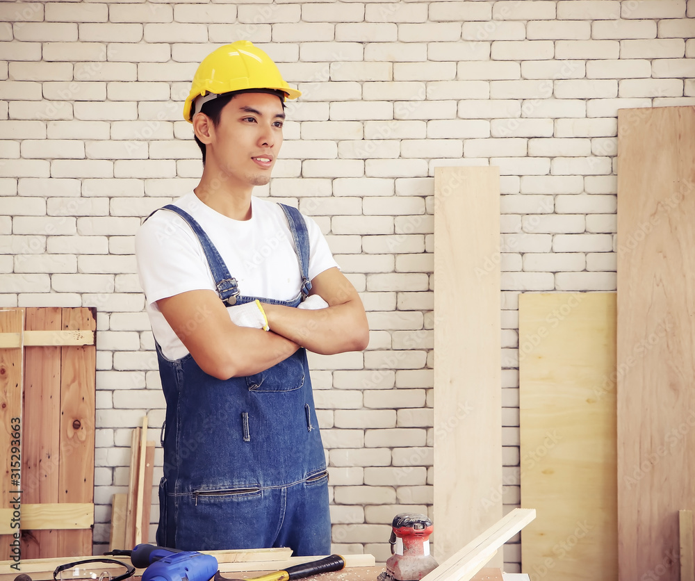handsome Asian repairman wearing yellow helmet standing and crossing his arms behind table with woodworking tools in workshop with white brick wall. handsome Asian repairman wearing yellow helmet standing and crossing his arms behind table with woodworking tools in workshop with white brick wall.