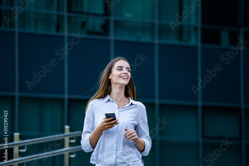 Happy smiling business woman talking on mobile smartphone in the street with office buildings in the background. Young woman with smartphone standing against street blurred building background.