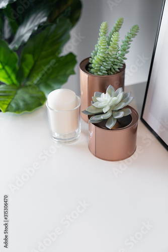 Bedroom working corner decorated with copper vase with artificial plant inside and white candle in glass on cream spray-painted working table with gray painted wall in the background /copy space