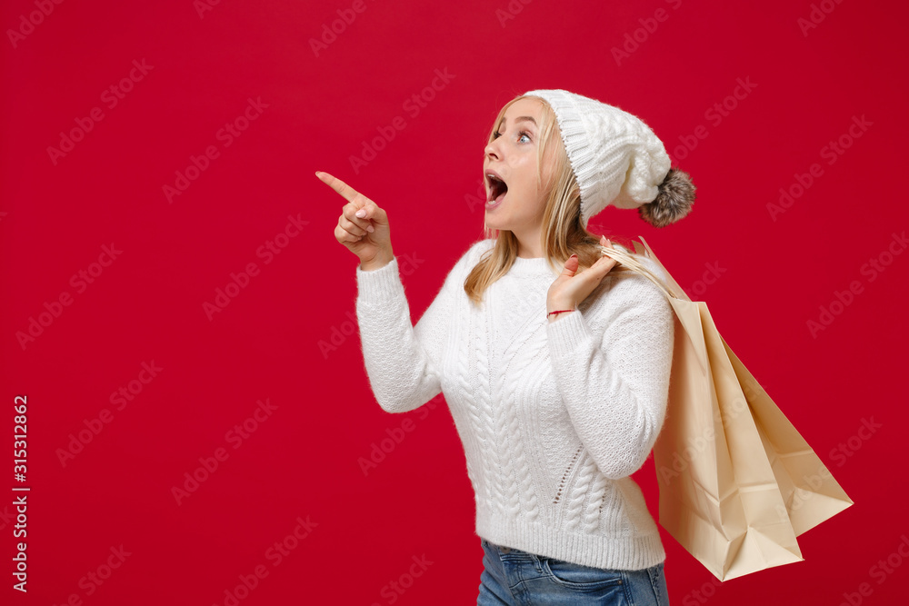 Amazed woman in sweater hat isolated on red background. Healthy fashion lifestyle, cold season concept. Mock up copy space. Hold package bag with purchases after shopping, pointing index finger aside. Amazed woman in sweater hat isolated on red background. Healthy fashion lifestyle, cold season concept. Mock up copy space. Hold package bag with purchases after shopping, pointing index finger aside.