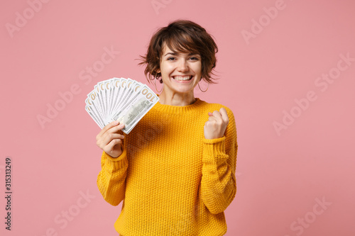 Joyful young brunette woman in yellow sweater posing isolated on pastel pink background. People lifestyle concept. Mock up copy space. Hold fan of cash money in dollar banknotes, doing winner gesture. photo