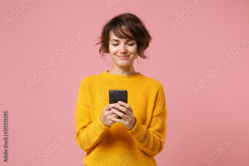 Smiling young brunette woman girl in yellow sweater posing isolated on pastel pink wall background studio portait. People lifestyle concept. Mock up copy space. Using mobile phone, typing sms message.