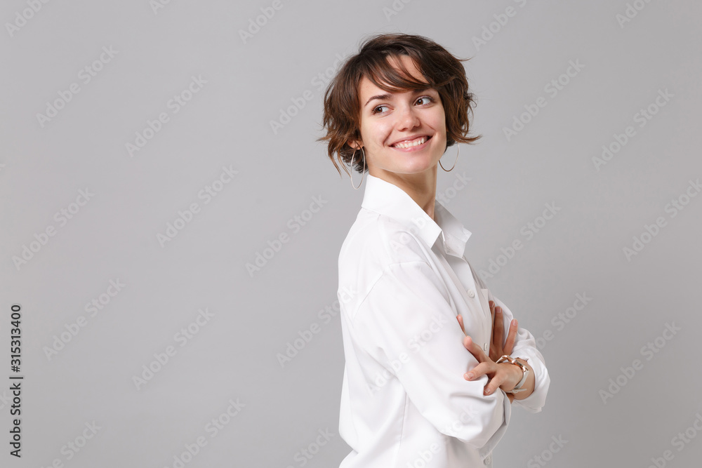 Side view of smiling young business woman in white shirt posing isolated on grey wall background. Achievement career wealth business concept. Mock up copy space. Holding hands crossed, looking aside. Side view of smiling young business woman in white shirt posing isolated on grey wall background. Achievement career wealth business concept. Mock up copy space. Holding hands crossed, looking aside.