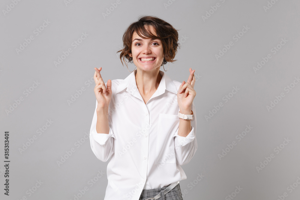 Smiling young business woman in white shirt isolated on grey background. Achievement career wealth business concept. Mock up copy space. Waiting for special moment keeping fingers crossed making wish. Smiling young business woman in white shirt isolated on grey background. Achievement career wealth business concept. Mock up copy space. Waiting for special moment keeping fingers crossed making wish.