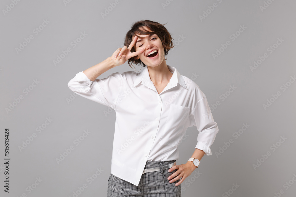 Cheerful young business woman in white shirt posing isolated on grey wall background studio portrait. Achievement career wealth business concept. Mock up copy space. Blinking, showing victory sign. Cheerful young business woman in white shirt posing isolated on grey wall background studio portrait. Achievement career wealth business concept. Mock up copy space. Blinking, showing victory sign.