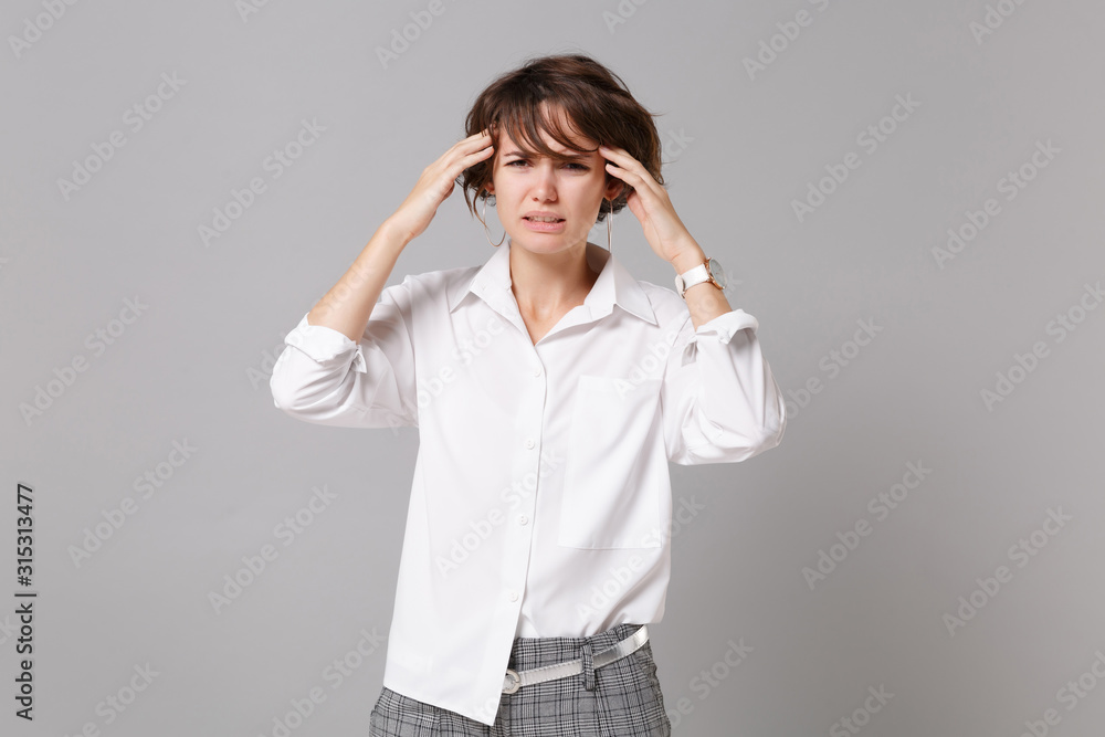 Tired sick young business woman in white shirt posing isolated on grey background studio portrait. Achievement career wealth business concept. Mock up copy space. Put hands on head, having headache. Tired sick young business woman in white shirt posing isolated on grey background studio portrait. Achievement career wealth business concept. Mock up copy space. Put hands on head, having headache.