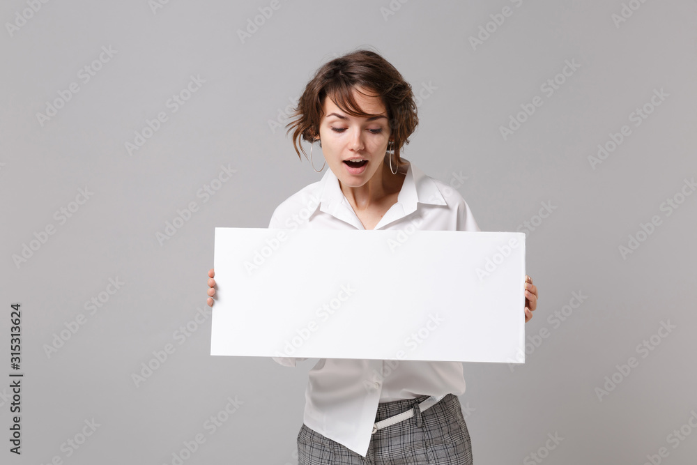 Curious funny young business woman in white shirt posing isolated on grey background. Achievement career wealth business concept. Mock up copy space. Hold white blank sign board with place for text. Curious funny young business woman in white shirt posing isolated on grey background. Achievement career wealth business concept. Mock up copy space. Hold white blank sign board with place for text.