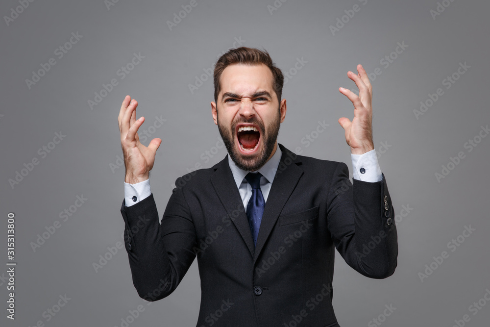 Angry mad young business man in classic black suit shirt tie posing isolated on grey background. Achievement career wealth business concept. Mock up copy space. Spreading hands, screaming swearing. Angry mad young business man in classic black suit shirt tie posing isolated on grey background. Achievement career wealth business concept. Mock up copy space. Spreading hands, screaming swearing.