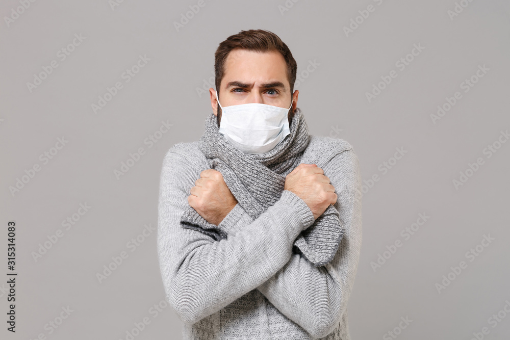 Young man in gray sweater, scarf isolated on grey background. Healthy lifestyle, ill sick disease treatment, cold season concept. Mock up copy space. Wearing sterile face mask, holding hands crossed. Young man in gray sweater, scarf isolated on grey background. Healthy lifestyle, ill sick disease treatment, cold season concept. Mock up copy space. Wearing sterile face mask, holding hands crossed.