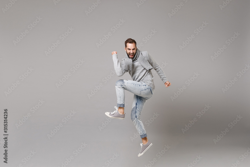 Cheerful young bearded man in gray sweater, scarf posing isolated on grey background, studio portrait. Healthy fashion lifestyle, cold season concept. Mock up copy space. Jumping doing winner gesture. Cheerful young bearded man in gray sweater, scarf posing isolated on grey background, studio portrait. Healthy fashion lifestyle, cold season concept. Mock up copy space. Jumping doing winner gesture.