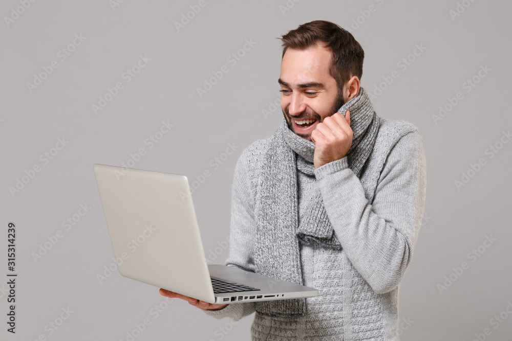 Funny young man in gray sweater, scarf isolated on grey background. Healthy lifestyle ill sick disease online treatment consulting, cold season concept. Mock up copy space. Hold laptop pc computer. Funny young man in gray sweater, scarf isolated on grey background. Healthy lifestyle ill sick disease online treatment consulting, cold season concept. Mock up copy space. Hold laptop pc computer.
