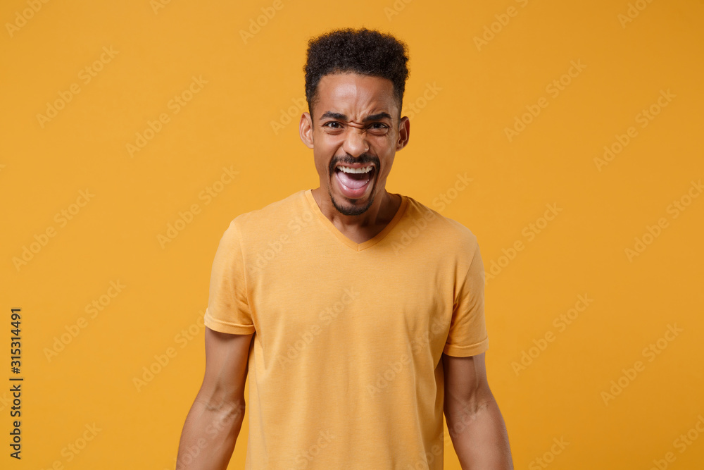 Shocked nervous young african american guy in casual t-shirt posing isolated on yellow orange wall background studio portrait. People sincere emotions lifestyle concept. Mock up copy space. Screaming. Shocked nervous young african american guy in casual t-shirt posing isolated on yellow orange wall background studio portrait. People sincere emotions lifestyle concept. Mock up copy space. Screaming.