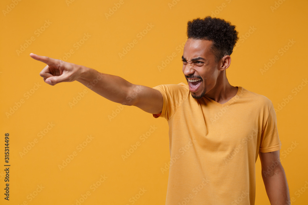 Angry young african american guy in casual t-shirt posing isolated on yellow orange background in studio. People lifestyle concept. Mock up copy space. Swearing screaming pointing index finger aside. Angry young african american guy in casual t-shirt posing isolated on yellow orange background in studio. People lifestyle concept. Mock up copy space. Swearing screaming pointing index finger aside.