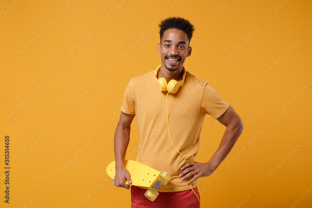 Smiling young african american guy in casual t-shirt, headphones posing isolated on yellow orange wall background studio portrait. People lifestyle concept. Mock up copy space. Holding skateboard. Smiling young african american guy in casual t-shirt, headphones posing isolated on yellow orange wall background studio portrait. People lifestyle concept. Mock up copy space. Holding skateboard.
