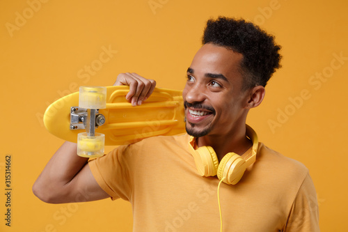 Smiling young african american guy in casual t-shirt, headphones posing isolated on yellow orange background in studio. People lifestyle concept. Mock up copy space. Holding skateboard, looking aside. photo