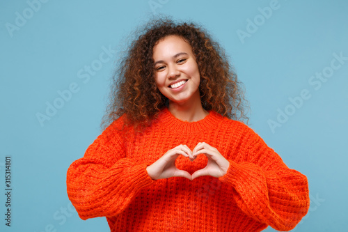 Smiling young african american girl in casual orange knitted clothes isolated on pastel blue background. People lifestyle concept. Mock up copy space. Showing shape heart with hands, heart-shape sign.