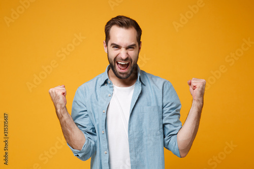 Joyful young bearded man in casual blue shirt posing isolated on yellow orange wall background, studio portrait. People sincere emotions lifestyle concept. Mock up copy space. Doing winner gesture. photo