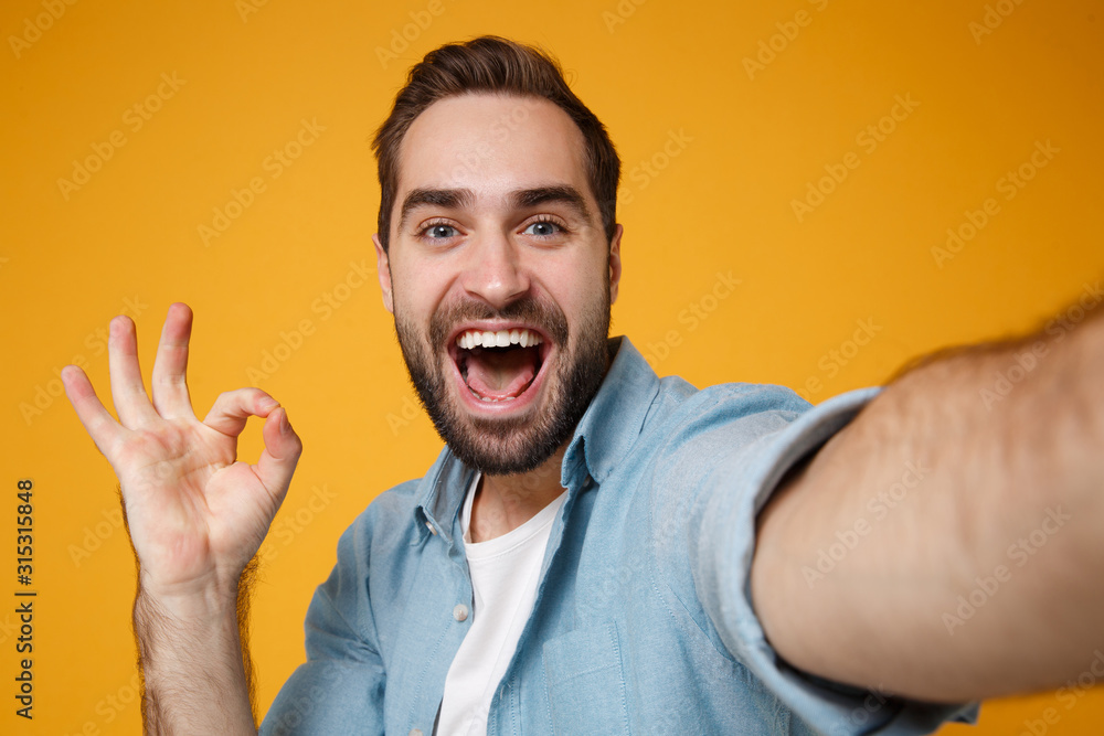 Close up of excited young man in casual blue shirt posing isolated on yellow orange wall background. People lifestyle concept. Mock up copy space. Doing selfie shot on mobile phone showing OK gesture.