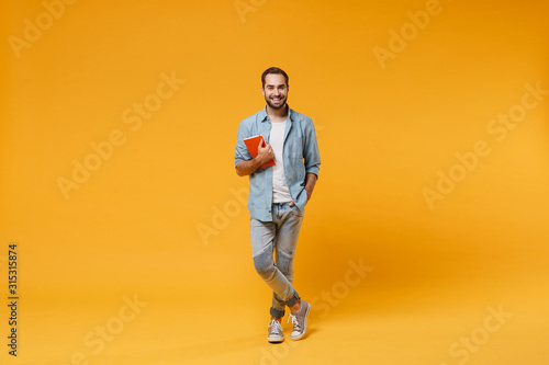 Smiling young student man in casual blue shirt posing isolated on yellow orange wall background studio portrait. People sincere emotions lifestyle concept. Mock up copy space. Holding red notebook.